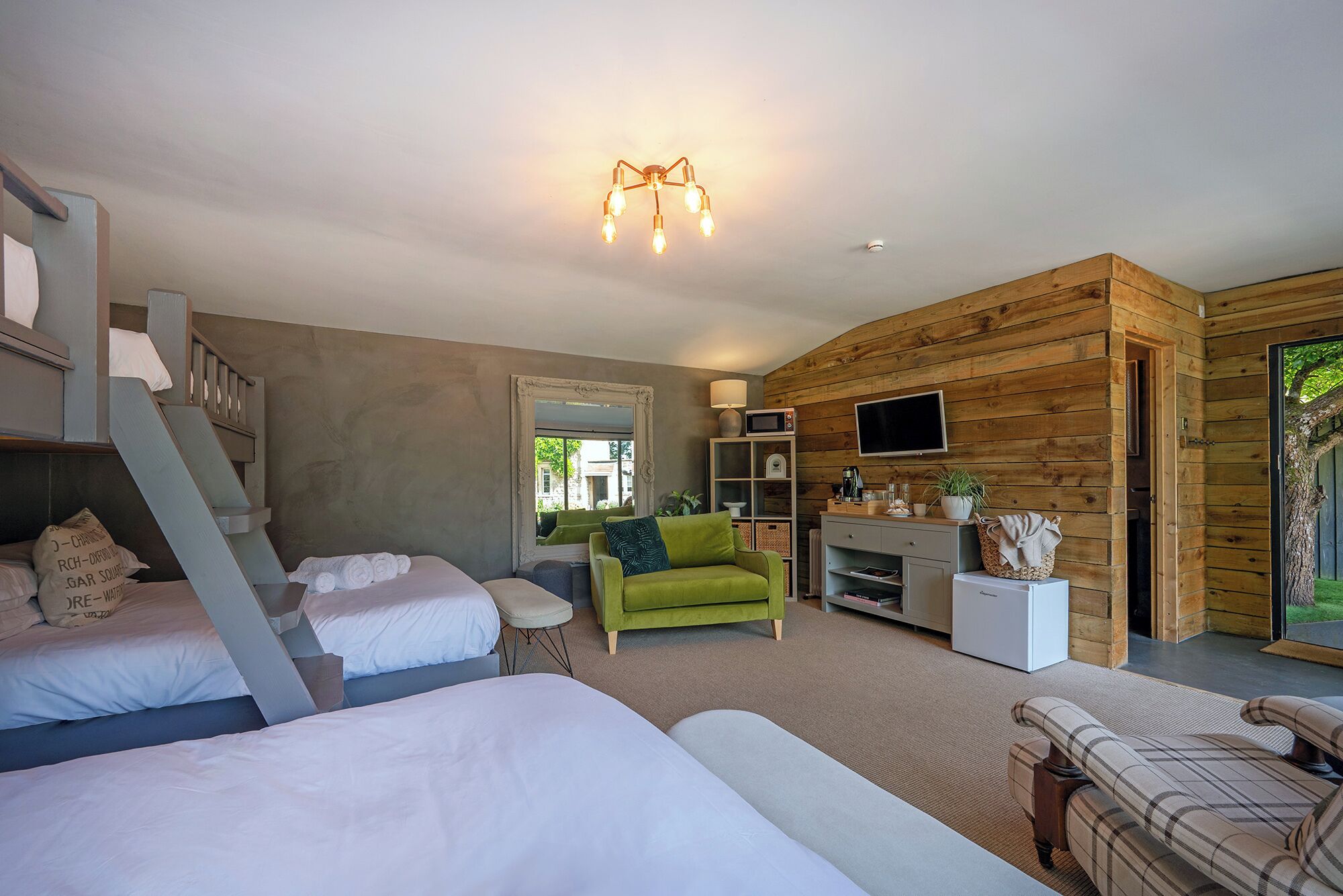 A cozy room with a bed, a green armchair, a sofa, a TV, and a wooden wall.