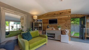 A modern living room with a green sofa, a large mirror, wooden panel walls, a flat-screen TV, and a small kitchen area. - Lavender Lodge  (Bath)