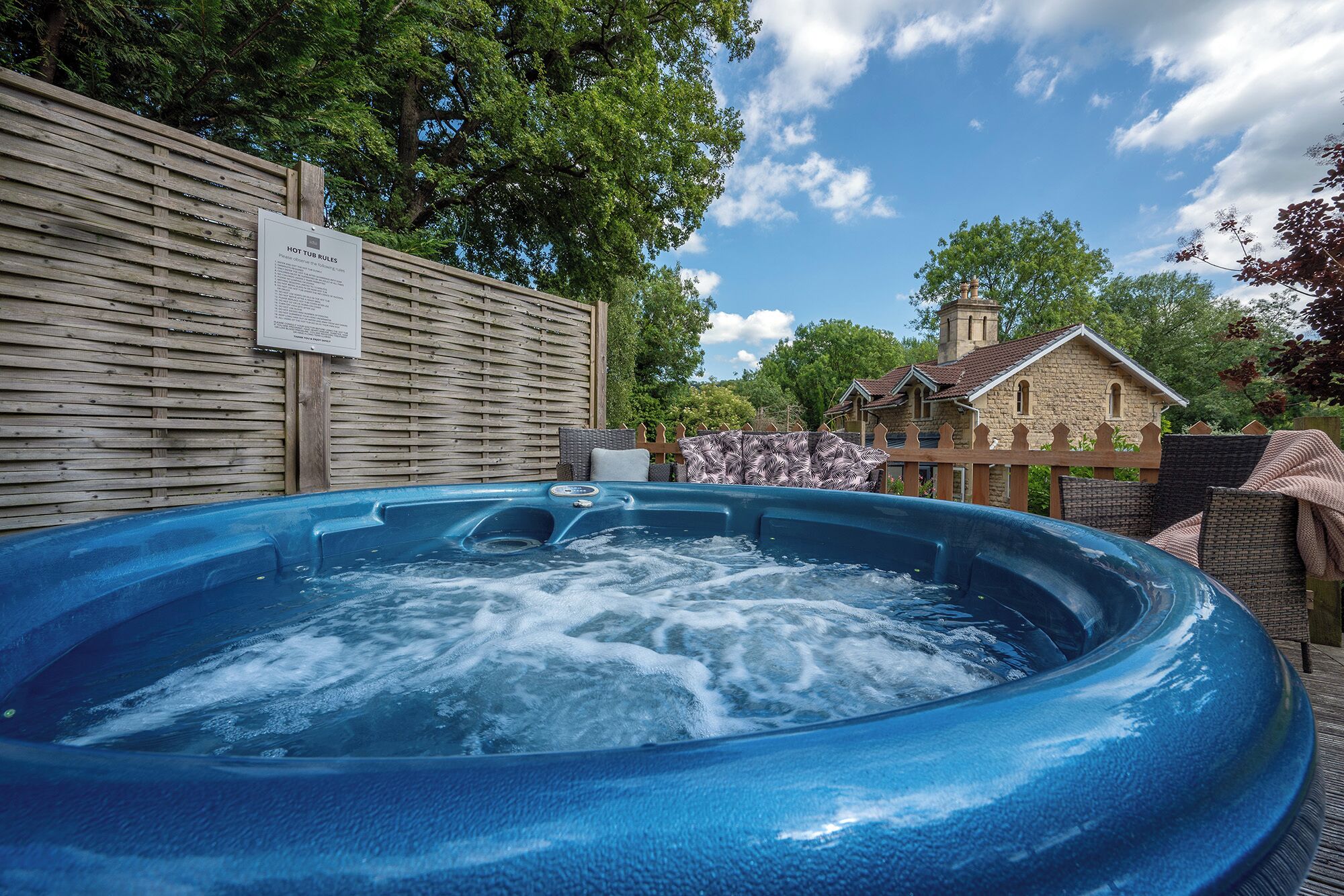 A hot tub with a wooden fence and a sign, surrounded by trees and a stone building.