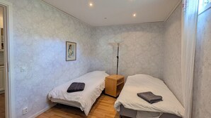 Een kleine slaapkamer met twee eenpersoonsbedden, een houten nachtkastje, een vloerlamp en een kamerkleed met dessin.