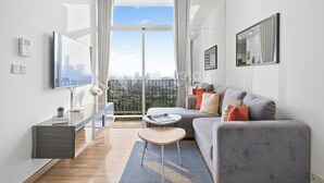 Ein modernes Wohnzimmer mit grauem Sofa, einem kleinen runden Tisch, einem Flachbildfernseher und einem Balkon mit Blick auf die Stadt.