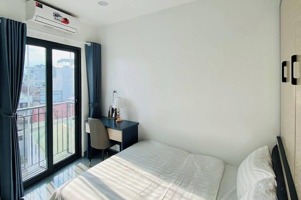 Une chambre d’hôtel avec un lit, un bureau et une chaise. Il y a une fenêtre avec des rideaux et un appareil de climatisation installé au-dessus.