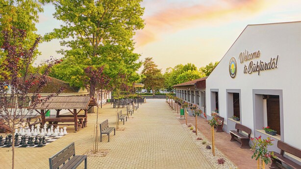 A paved walkway with benches and chessboards, a white building with a sign reading "Welcome to Büfflirdö!", and trees lining the path. - Park Hotel Bükfürdő (Bükfürdo)