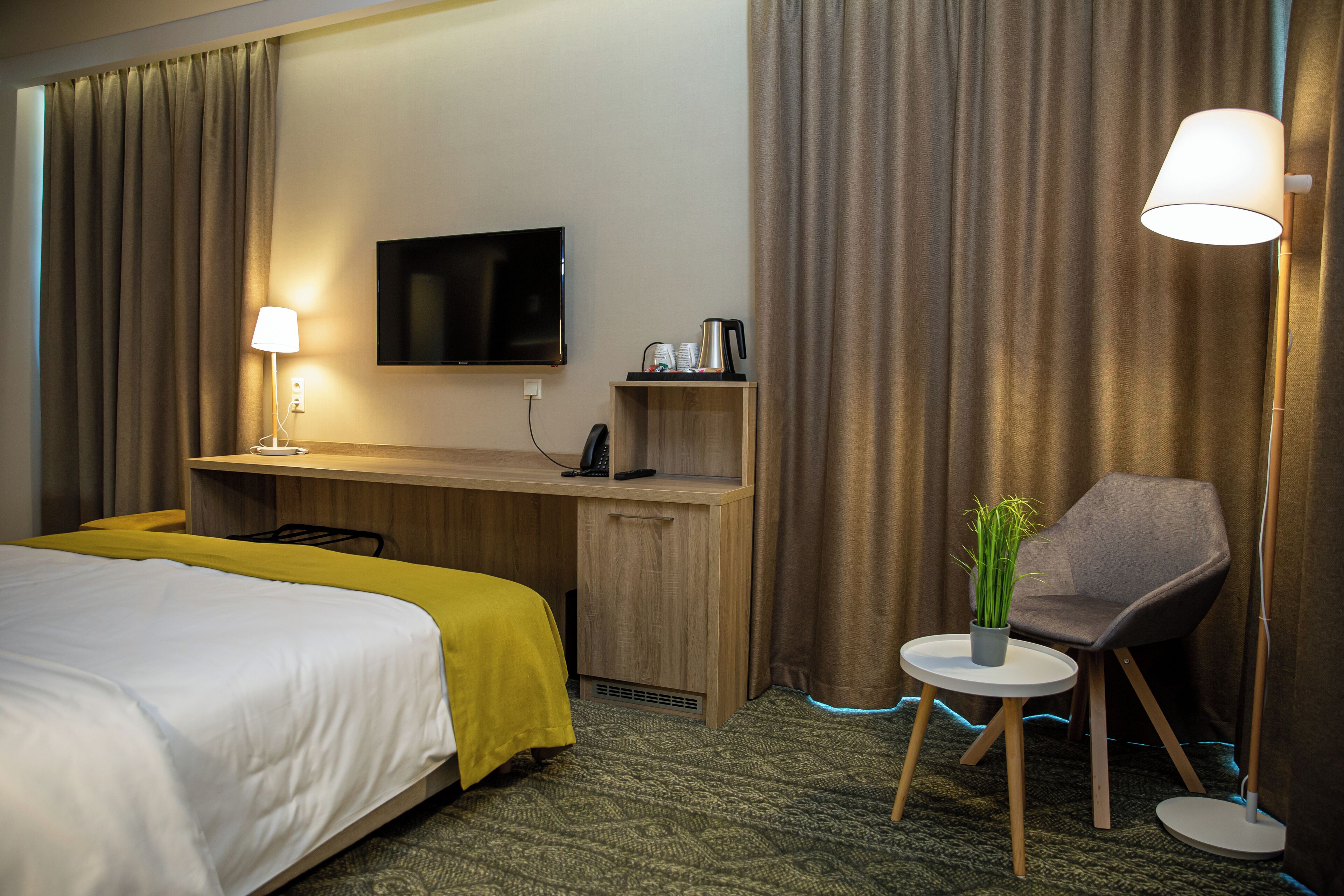 Ein Hotelzimmer mit Bett, Schreibtisch, Stuhl, Fernseher, Lampe und Topfpflanze.
