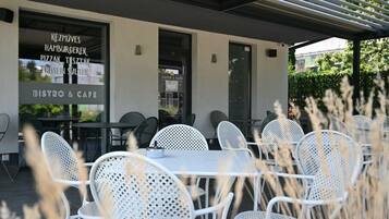 An outdoor cafe with white metal chairs and tables, a glass wall with menu options, and a view of greenery.