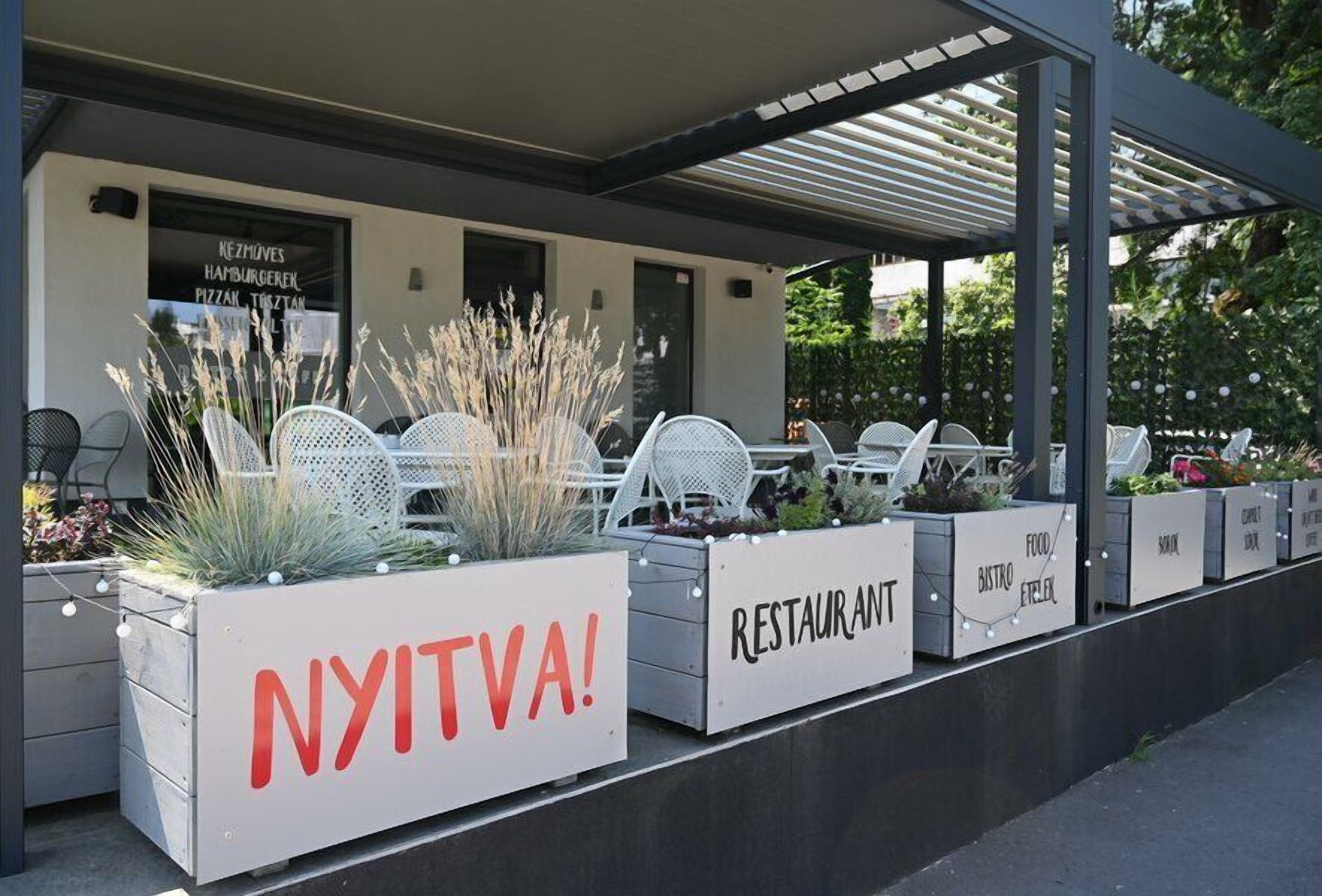 Ulkoterassi, jossa on valkoisia tuoleja ja istutusastioita, katettu patio ja kyltti, jossa lukee: ”AVOINNA!”.