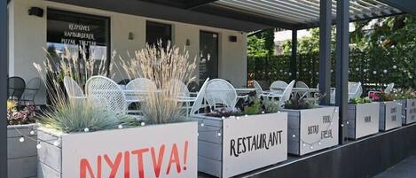 Ulkoterassi, jossa on valkoisia tuoleja ja istutusastioita, katettu patio ja kyltti, jossa lukee: ”AVOINNA!”.