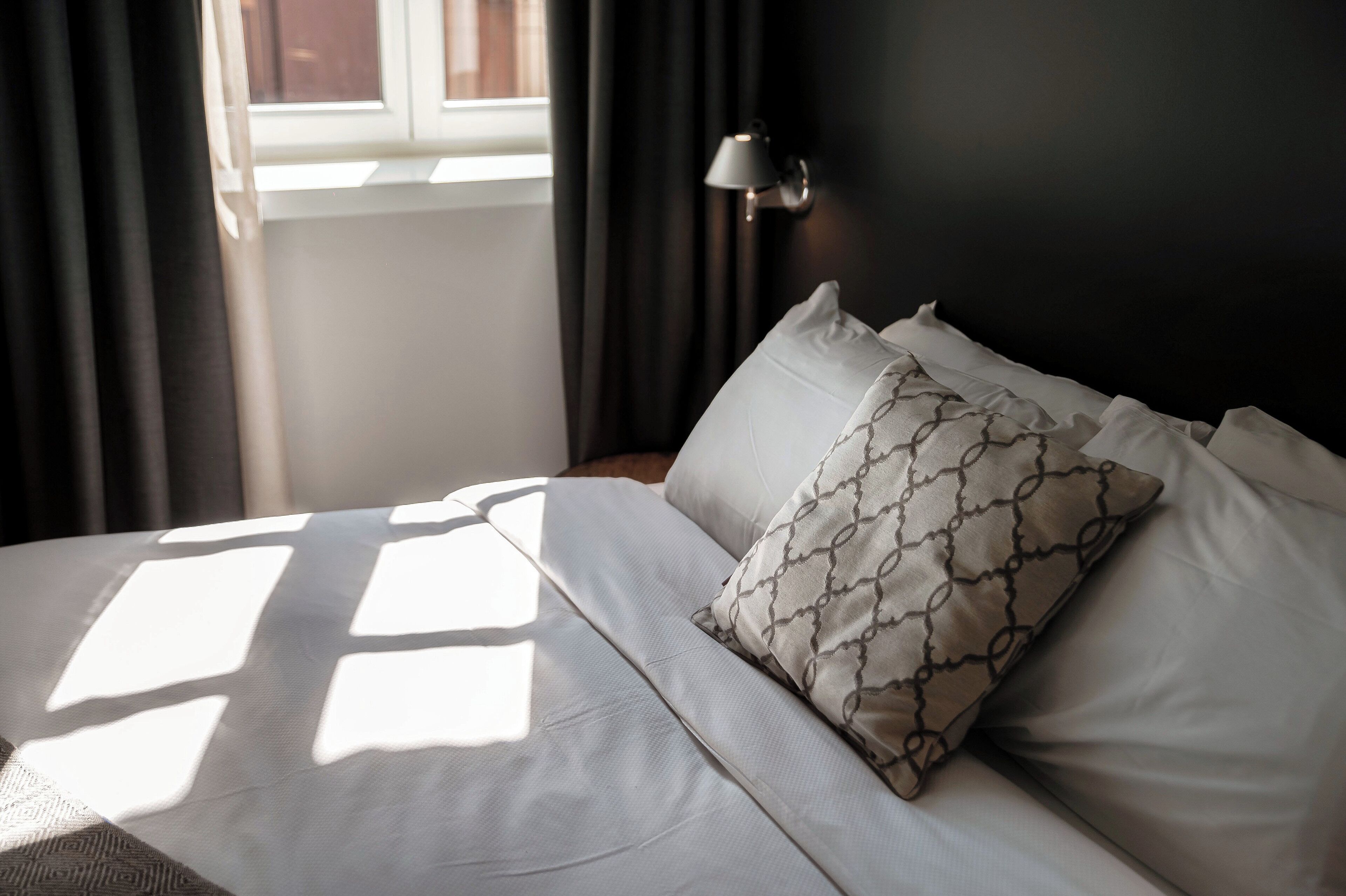 A neatly made bed with white linens and patterned pillows, a bedside lamp, and a window with curtains.