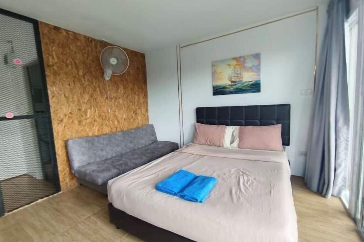 A bedroom with a bed, a grey sofa, a wooden accent wall, a painting, and a window with curtains.