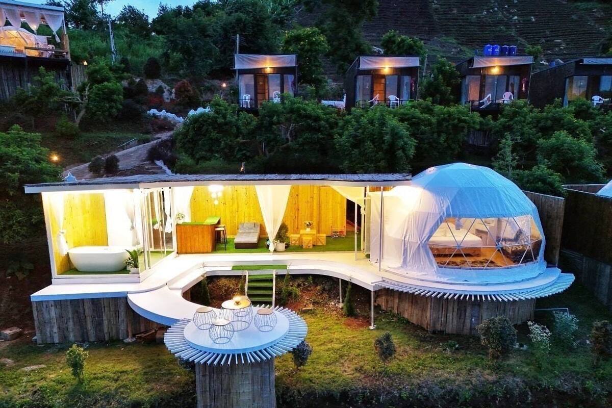 Una casa moderna i ecològica amb una estructura de cúpula central, envoltada de terrasses de fusta i un petit jardí.