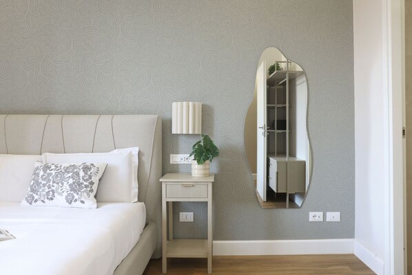 A bedroom with a bed, bedside table, lamp, mirror, and a potted plant.