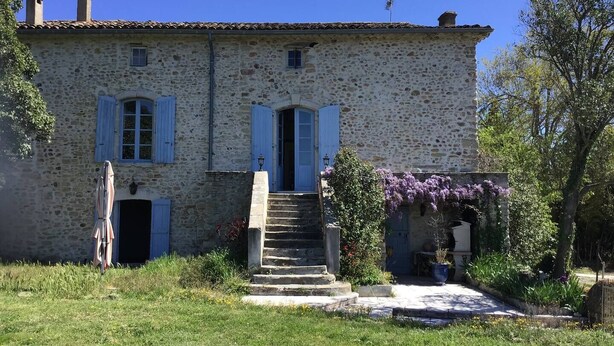 Un edificio de piedra con una escalera central, dos puertas con contraventanas y un jardín con flores y plantas.