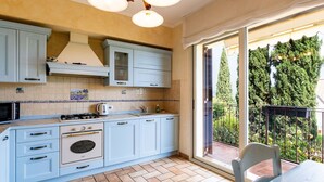 A kitchen with light blue cabinets, a gas stove, and a large window leading to a balcony with a view of trees and a railing. - Nice apartment in villa for 5 people with A/C, WIFI, pool, terrace and pets allowed (Borghetto Santo Spirito)