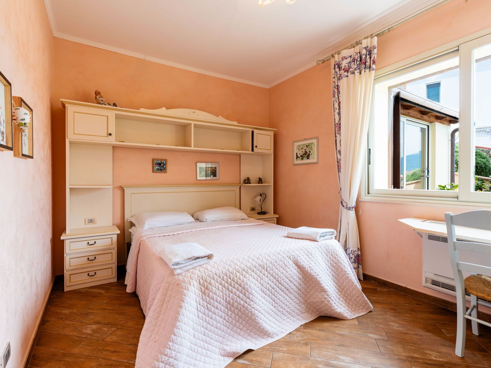Une chambre à coucher avec un lit, un bureau et une chaise. Il y a des étagères au-dessus du lit et une fenêtre avec des rideaux.