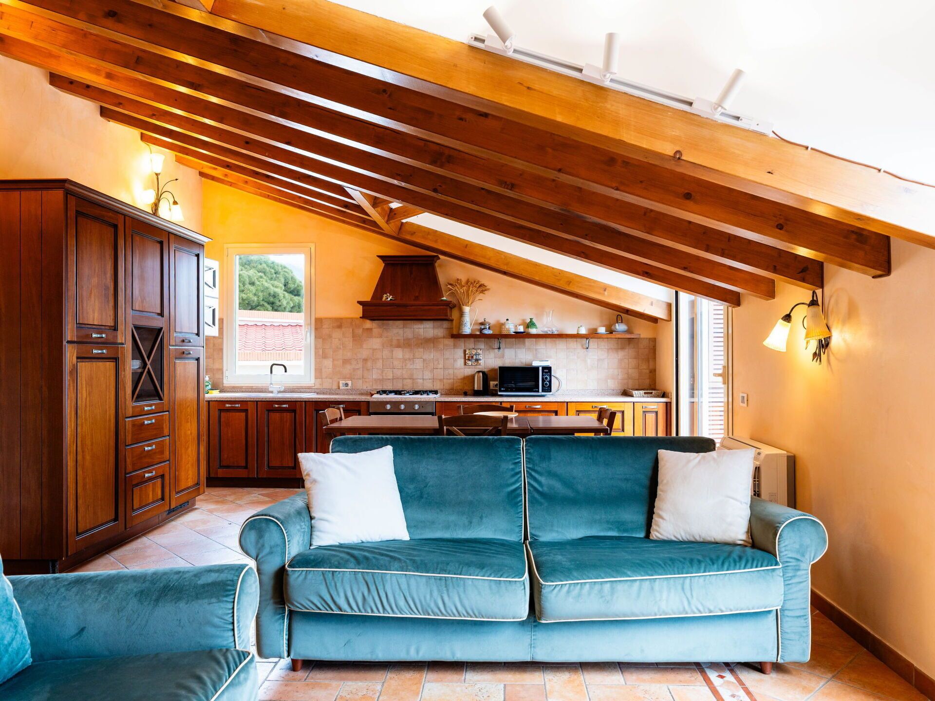 Un'accogliente zona giorno con un divano color petrolio, mobili contenitori in legno e una cucina con vista dalla finestra.