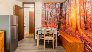 A compact kitchen with a refrigerator, a dining table set for two, and a large autumn-themed wall mural. - Nice apartment for 6 guests with WIFI, TV, patio and panoramic view (Mezzana)