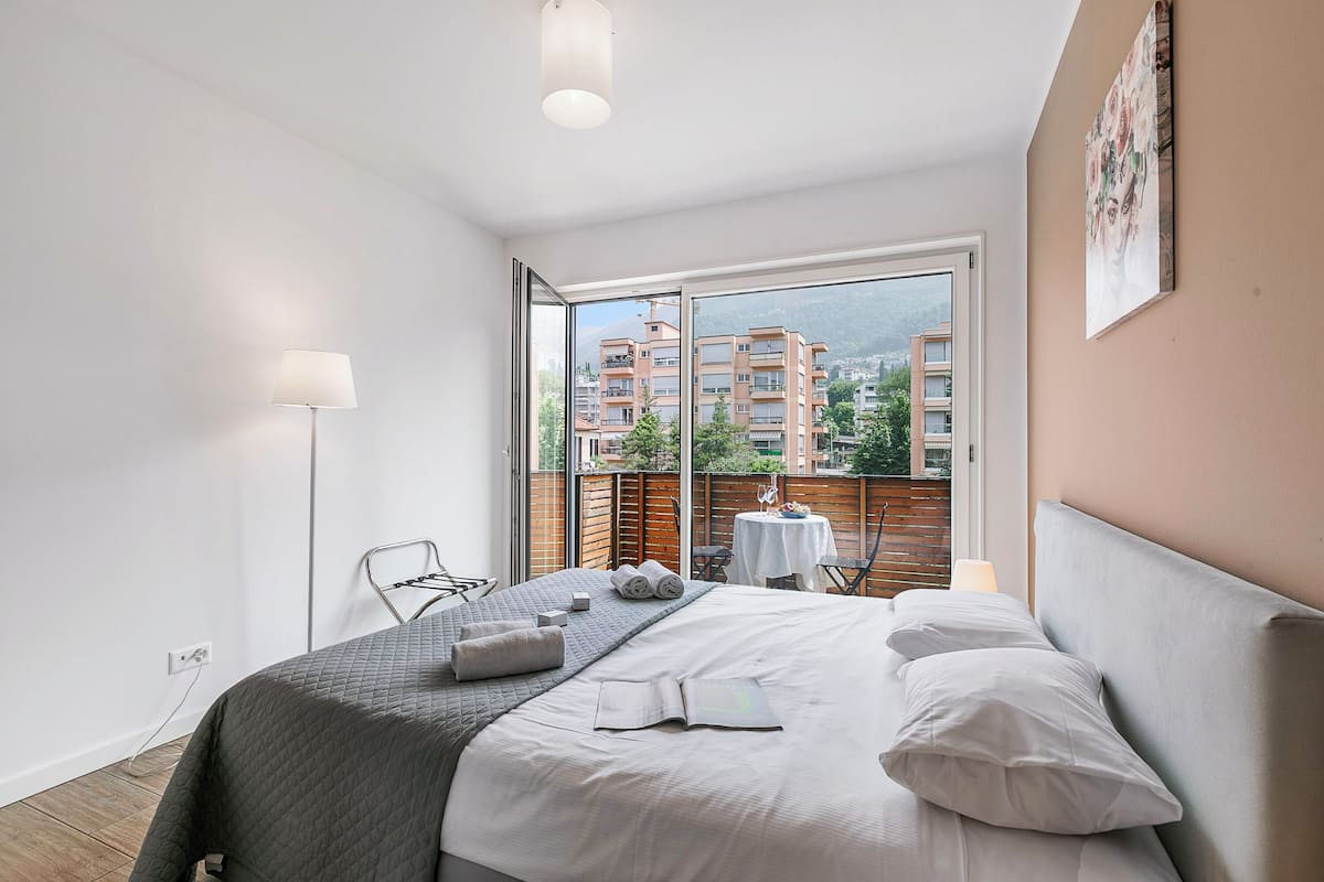A modern bedroom with a large bed, a view of buildings and mountains through a window, a small table with a chair, and a lamp.