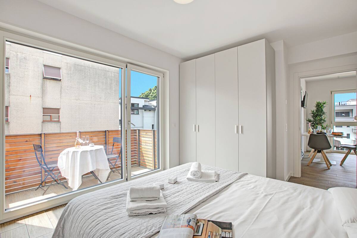 A modern bedroom with a large bed, a sliding glass door to a balcony, and a view of the city.