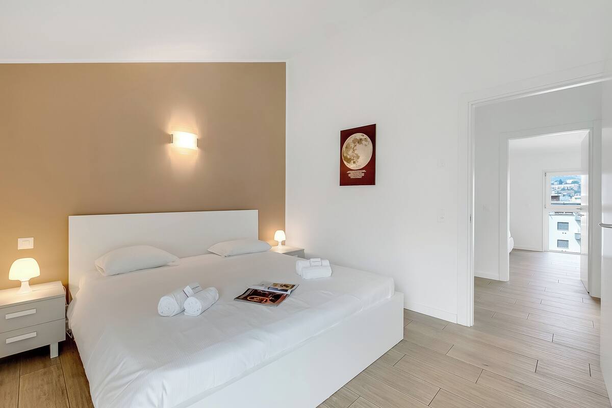 A neatly made bed with white linens, a magazine, and rolled towels on a wooden floor.