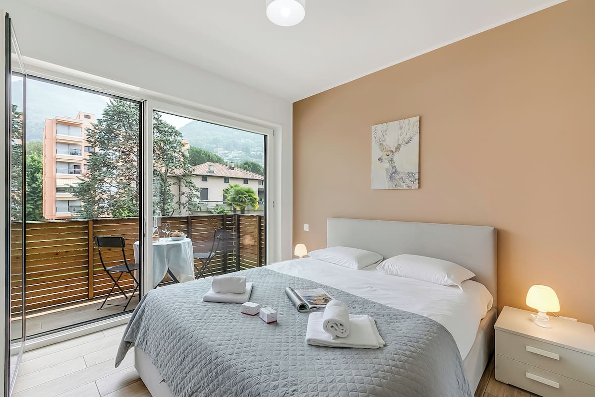 A modern hotel room with a large bed, a bedside table with a lamp, a view of the city through large windows, and a balcony with a chair and table.