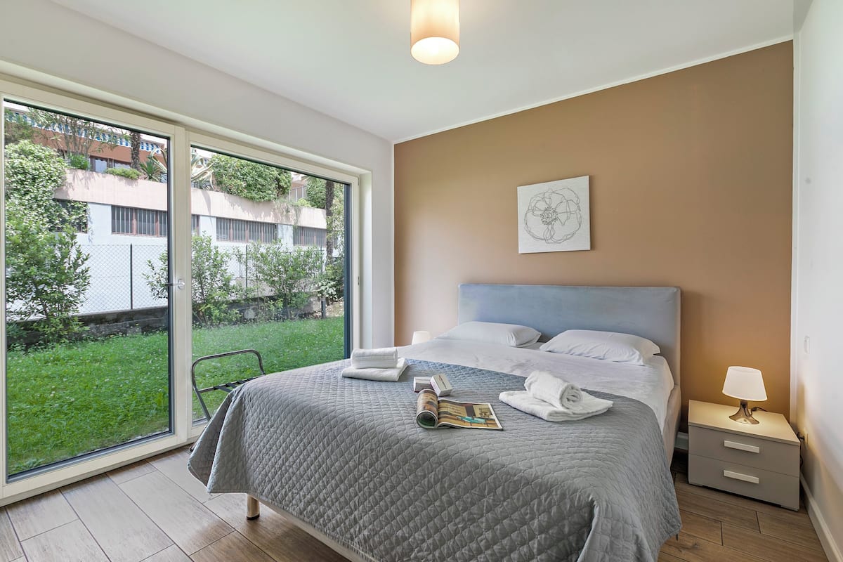 A modern bedroom with a large bed, a bedside table with a lamp, and a view of a green garden through a sliding door.