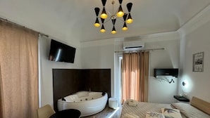 Habitación de hotel con una cama, una bañera de hidromasaje redonda, un televisor, un candelabro y una pequeña mesa con sillas.