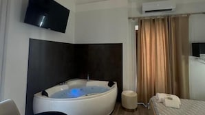 Una habitación de hotel moderna con una bañera grande y curva, un televisor de pantalla plana montado en la pared, una lámpara de techo y una cama con ropa de cama impecable.