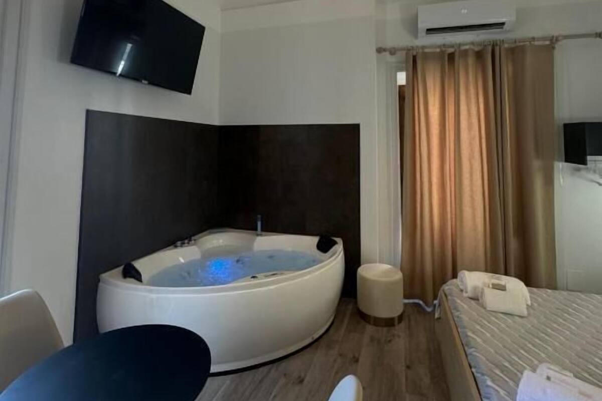 Una habitación de hotel moderna con una bañera grande y curva, un televisor de pantalla plana montado en la pared, una lámpara de techo y una cama con ropa de cama impecable.