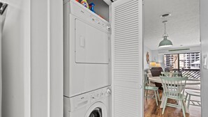 A laundry room with stacked washers and dryers, a dining area with a table and chairs, and a view of buildings through the window. - Relax, Explore, and Make Memories at Shipwatch Pointe I 216 F (Myrtle Beach)
