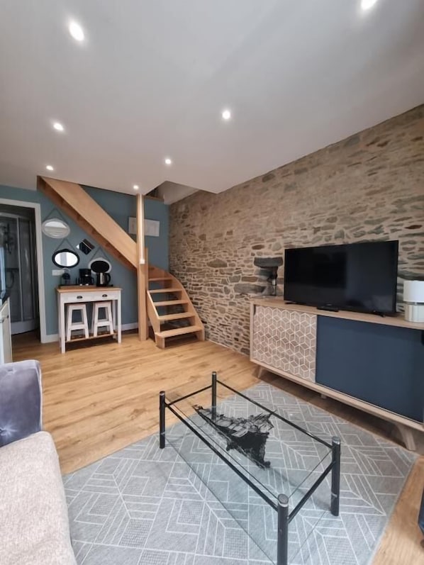 Un salon moderne avec un mur en pierre, un escalier en bois, une table basse en verre et un téléviseur posé sur un meuble.