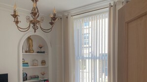A living room with a white sofa, a chandelier, a bookshelf with decorative items, and a window with vertical blinds.