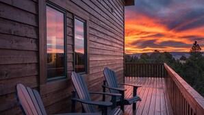 A wooden deck with Adirondack chairs, a wooden railing, and a view of a sunset over a forested landscape.