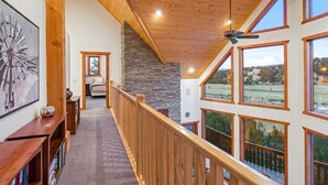 A spacious interior with a high ceiling, wooden beams, a stone fireplace, and large windows offering views of the outdoors.