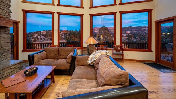 A spacious living room with a large sectional sofa, a wooden coffee table, and a stone fireplace. Large windows offer a view of a mountainous landscape.