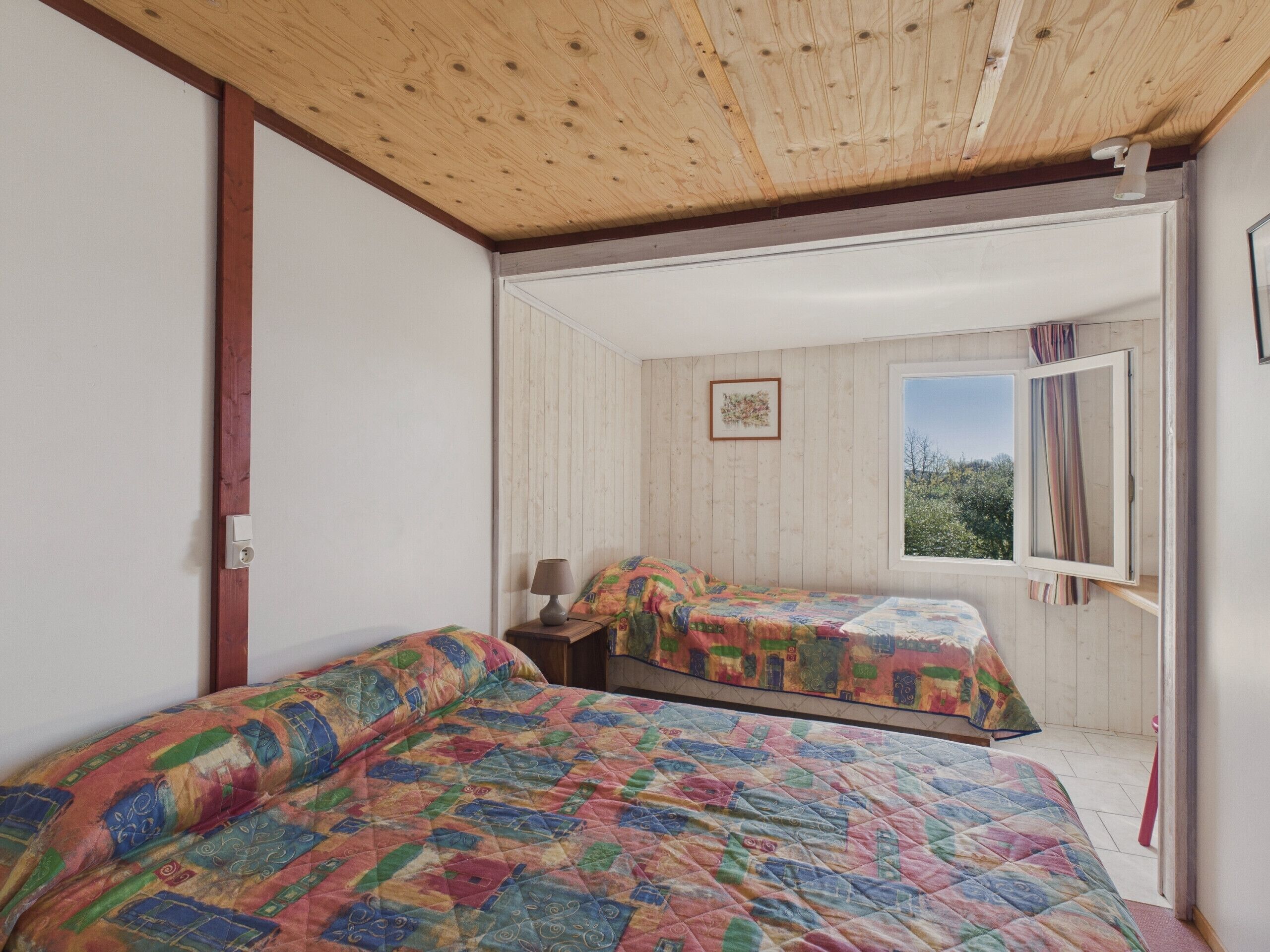 Una habitación con dos camas, techo de madera, una ventana con cortinas y una mesita con lámpara.