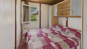 A bedroom with a wooden ceiling, a window with curtains, a bed with a patterned quilt, and built-in wooden cabinets. - Chalet La Brousse No. 2 – sleeps 5 – with pool (Salignac-Eyvigues)