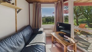A cozy living room with a leather sofa, a wooden coffee table, and a TV. There is a window with curtains and a view of a garden. - Chalet La Brousse No. 2 – sleeps 5 – with pool (Salignac-Eyvigues)