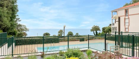 A pool area with a view of the sea, surrounded by a green fence and various plants.