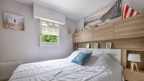 A neatly made bed with white bedding and a blue pillow, a wooden nightstand with a lamp, and a window with a view of greenery. - Apart 100m from the beach - swimming pool for 4 (Pornic)