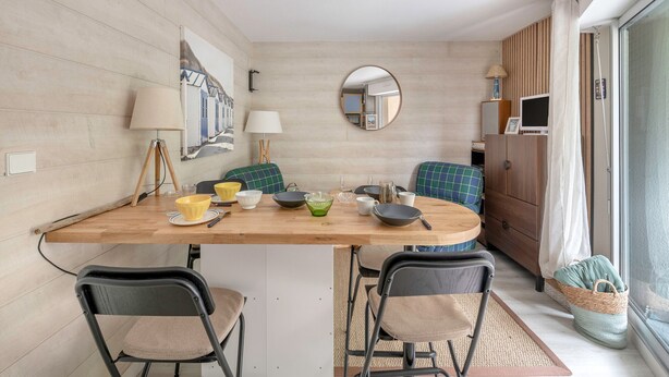 A compact dining area with a wooden table, four chairs, and various bowls and cups. A mirror hangs on the wall, and a window with curtains is visible. - Apart 100m from the beach - swimming pool for 4 (Pornic)