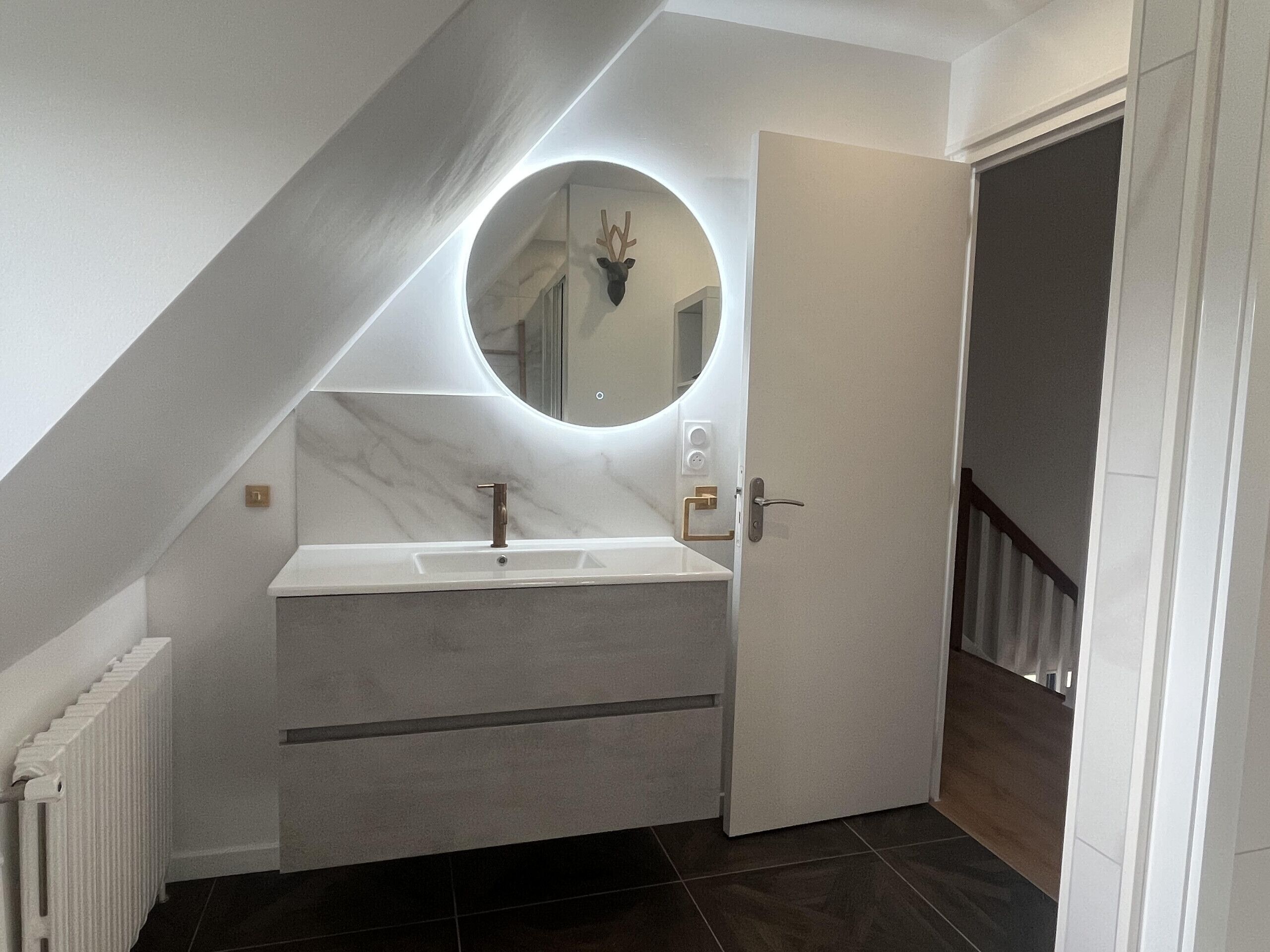 Et moderne badeværelse med et hvidt toiletbord, et rundt spejl og en dør til en trappe.