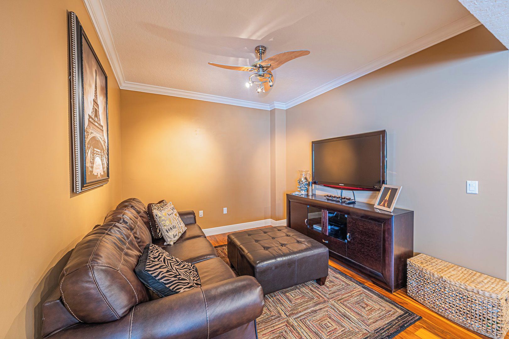 A living room with a brown leather sofa, a wooden coffee table, a flat-screen TV on a dark wooden stand, and a ceiling fan.