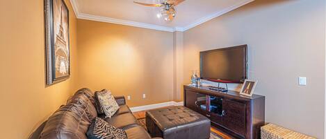 A living room with a brown leather sofa, a wooden coffee table, a flat-screen TV on a dark wooden stand, and a ceiling fan.