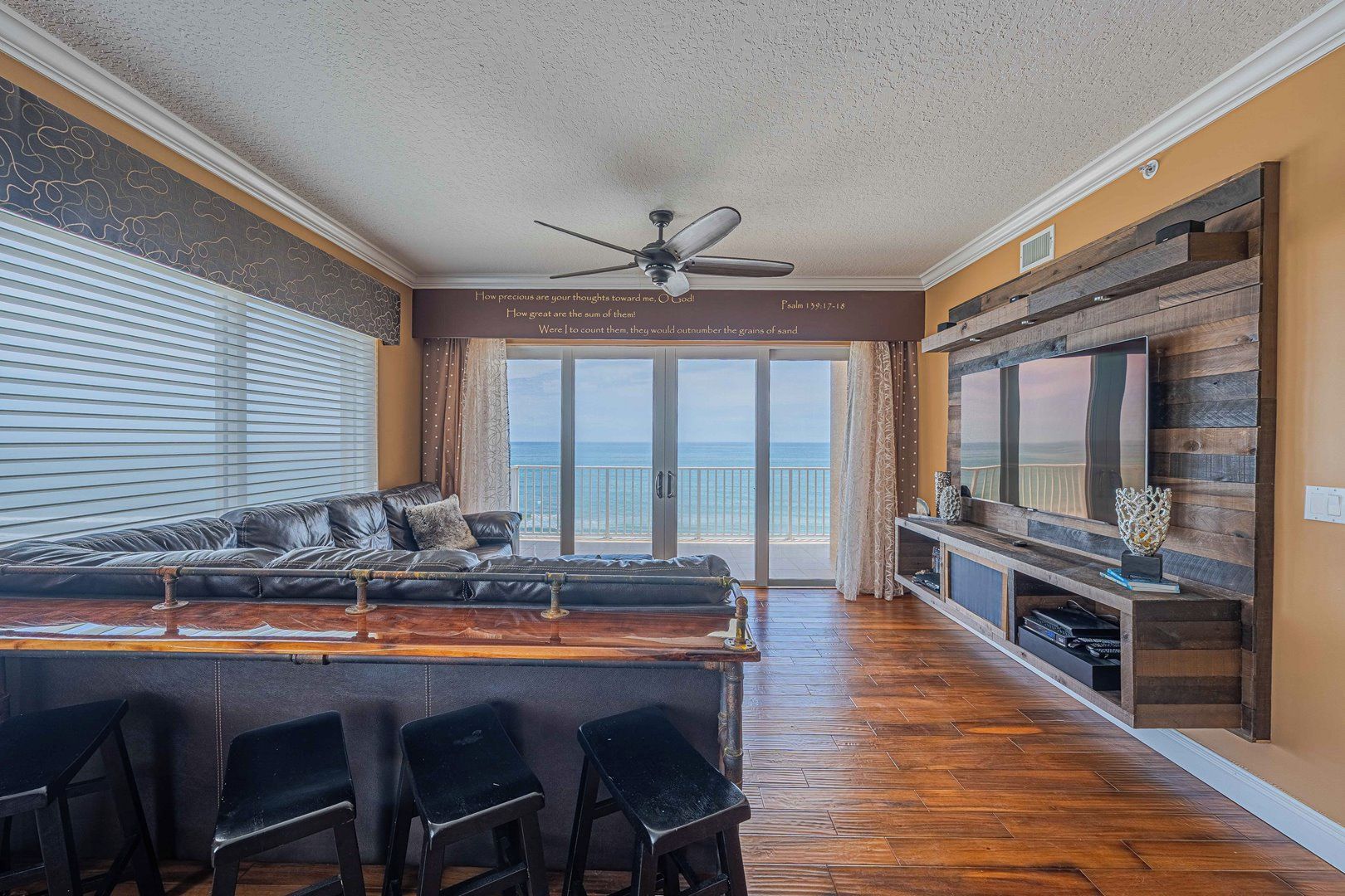 A spacious living room with a large sofa, a dining area with bar stools, a ceiling fan, and a large TV on a wooden wall.