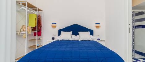 Une chambre à coucher avec un grand lit, des murs blancs, une tête de lit bleue et une armoire où sont suspendus des vêtements.