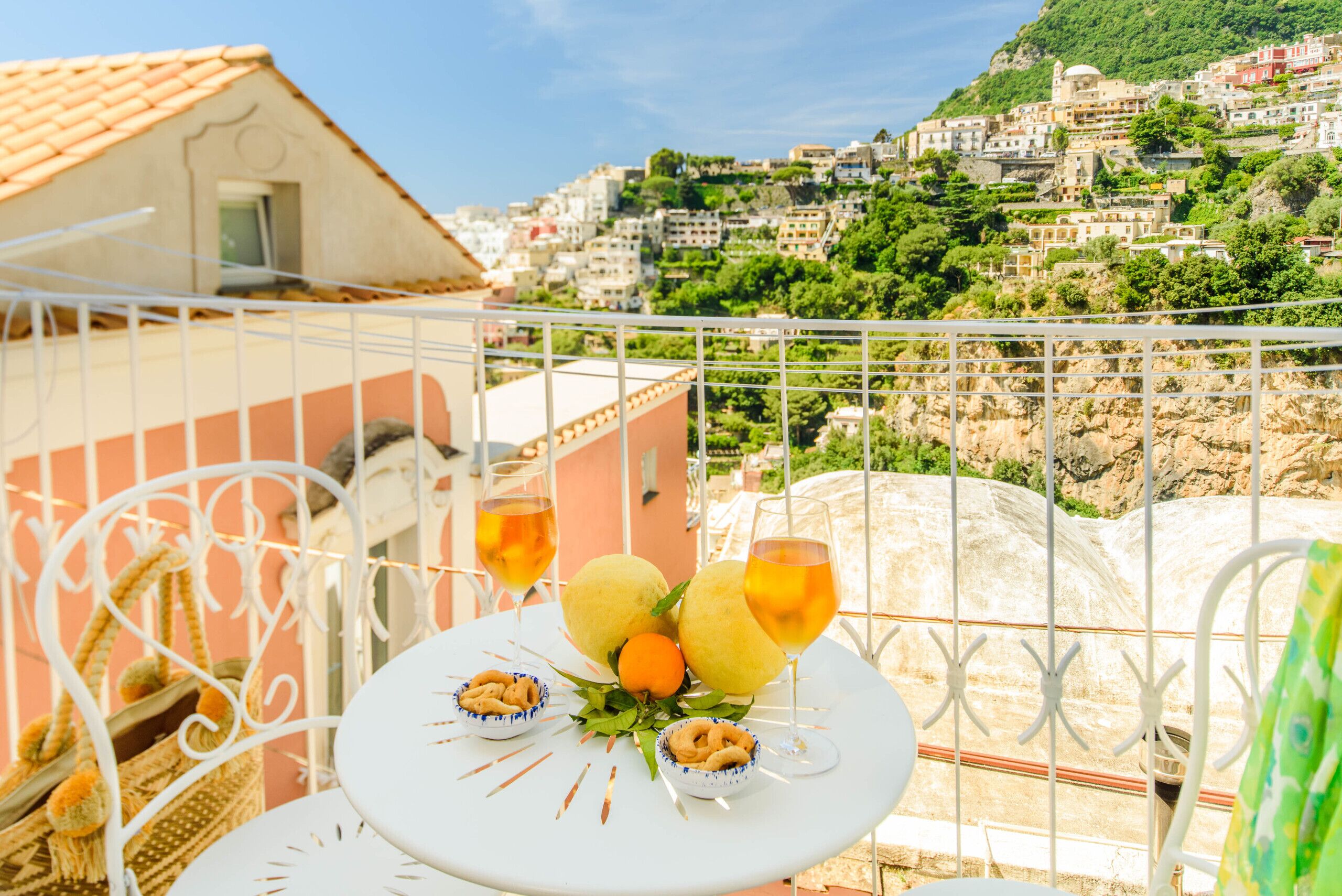 Un balcone con un tavolo apparecchiato per due persone, con un bicchiere di vino, una ciotola di frutta secca e della frutta. Sullo sfondo, si vede una collina con edifici.