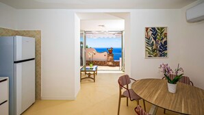 Una cocina con refrigerador, una zona de comedor con mesa y sillas, y vista al mar a través de una puerta abierta.