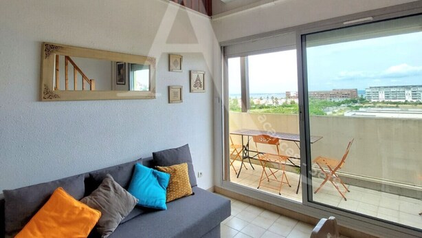 A modern living room with a grey sofa, colorful cushions, a wooden staircase, and a large sliding glass door leading to an outdoor balcony with a table and chairs.
