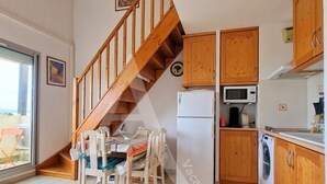A kitchen with wooden cabinets, a refrigerator, a microwave, and a washing machine. There is a dining table with chairs and a staircase leading to another level.