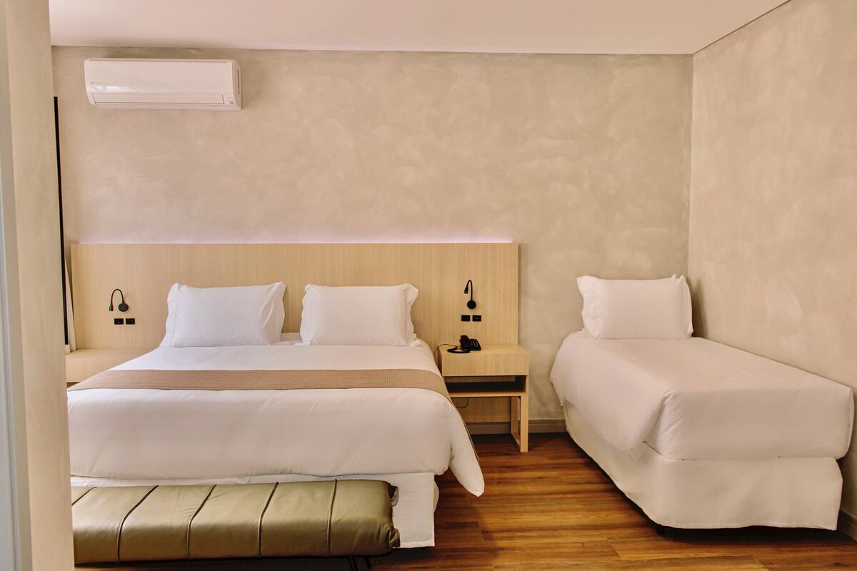 Hotelkamer met een groot bed, een kleiner bed, een bankje, een nachtkastje met een telefoon en een airconditioningsysteem aan de muur.